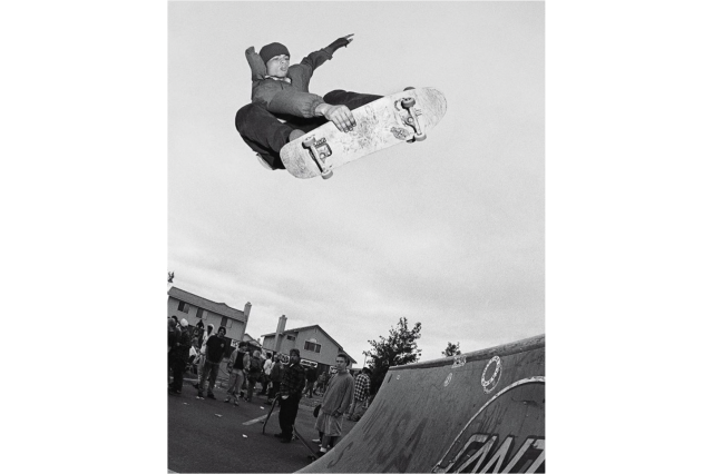 Black and white photo, view from below, of a skateboarder grabbing onto his board in midair above a skate ramp